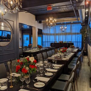 An opulent dining room with a long table set for a corporate event at Brix & Mortar, featuring dark chairs, white tablecloths, and vibrant red rose centerpieces. Crystal glassware sparkles under the warm light of two grand chandeliers, with sheer curtains in the background softening the daylight. Brix & Mortar is your destination for corporate events in Yaletown, Vancouver.