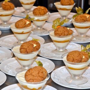 A collection of dessert glasses, each filled with alternating layers of white cream and caramel, topped with a large ginger snap cookie and a fresh mint leaf, all arranged on lace paper doilies for a sophisticated presentation which can be used for events or weddings at Brix & Mortar restaurant in Yaletown.